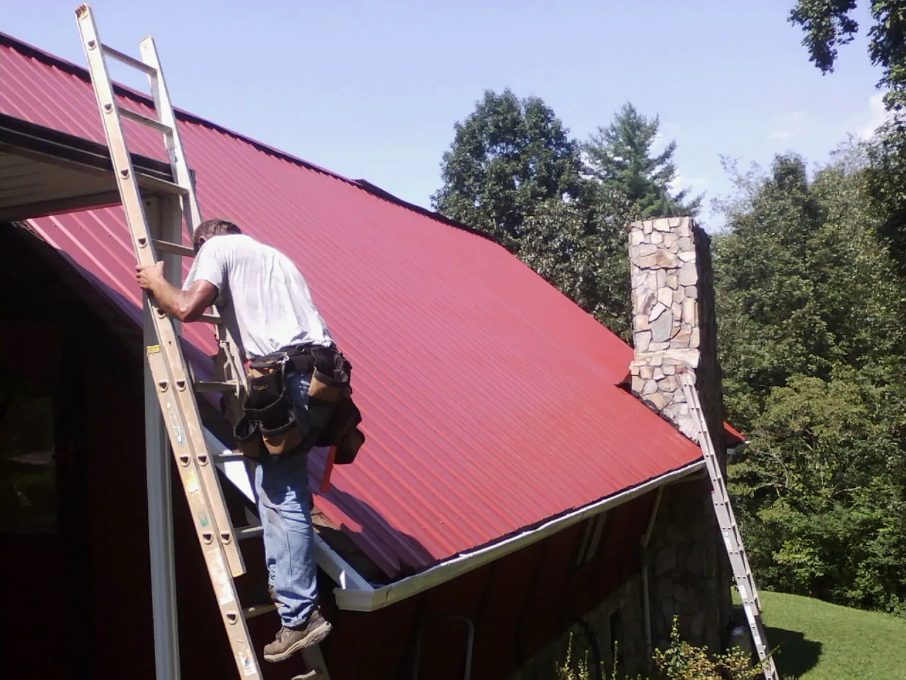 Roofer on a ladder beside a red metal roof and stone chimney — Soffit & Fascia Repair by Roofing Brothers in Downingtown